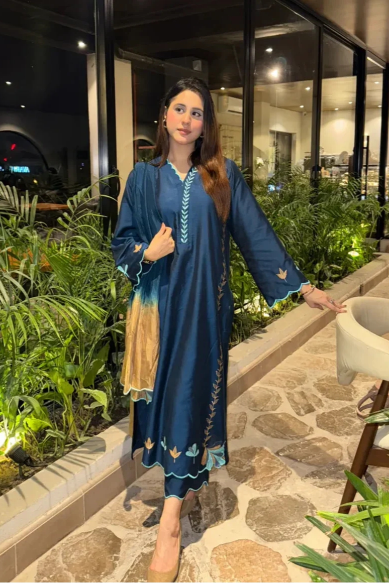 Woman in a blue traditional outfit standing in an outdoor setting with plants and a building in the background.