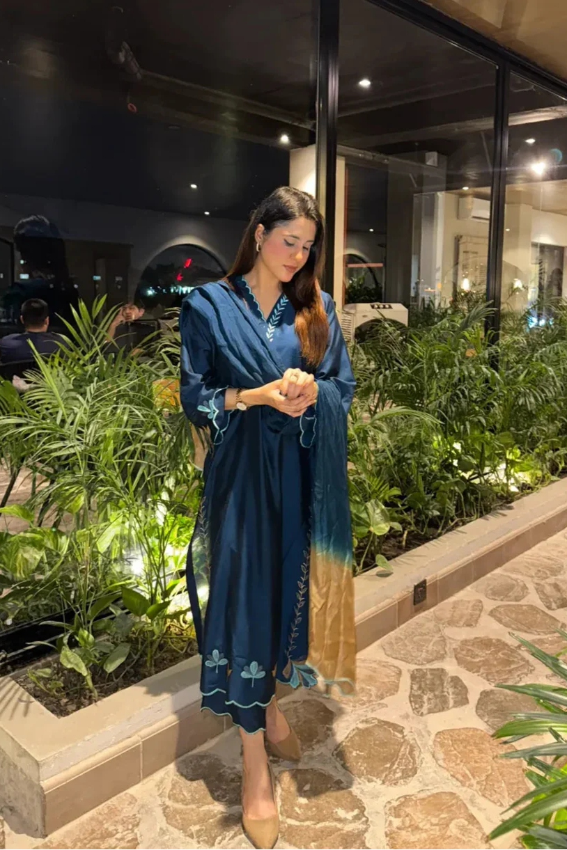 Woman in a blue dress standing in front of a glass wall with plants inside.