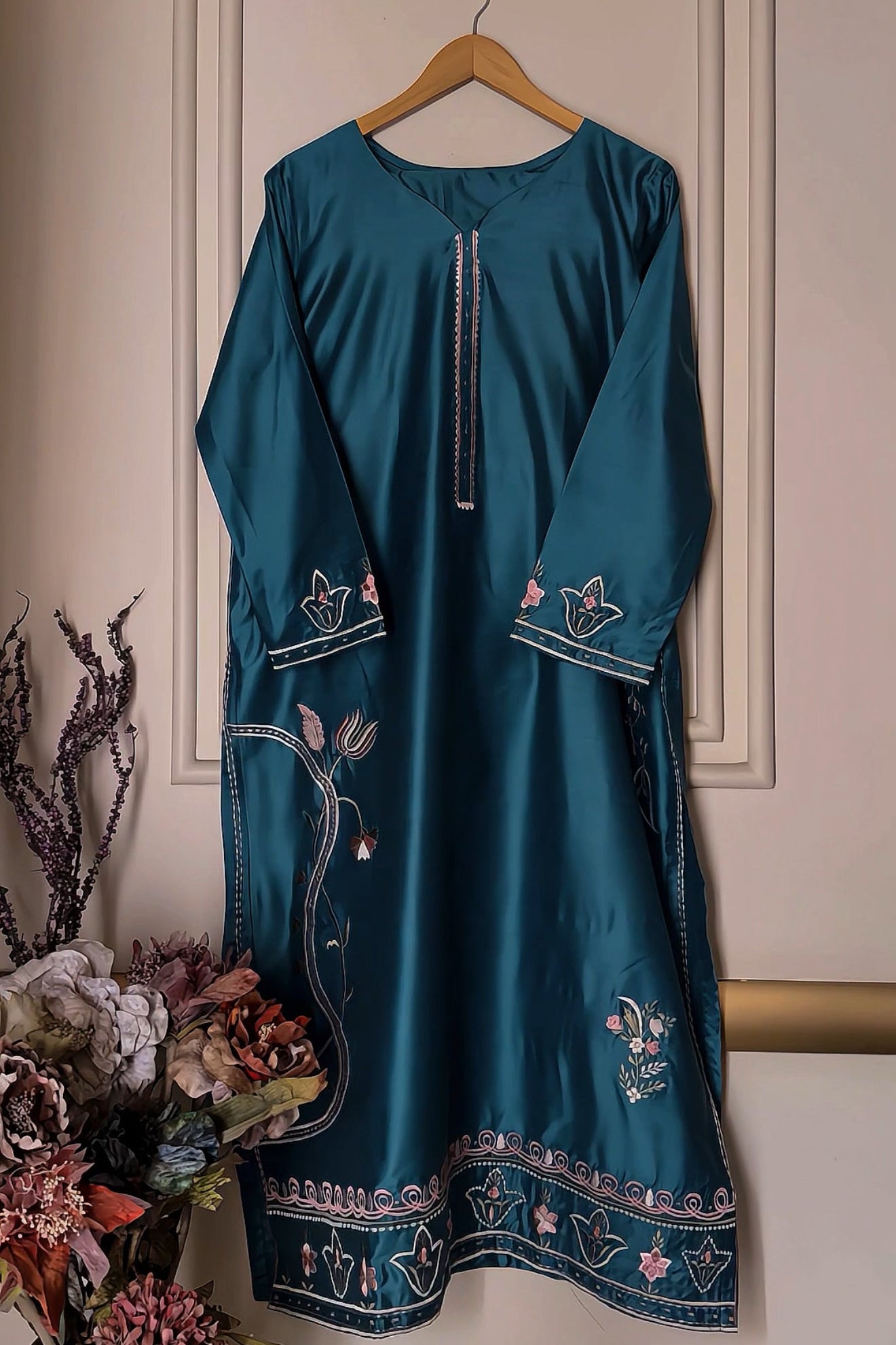 Teal dress with floral embroidery on a hanger against a neutral wall.